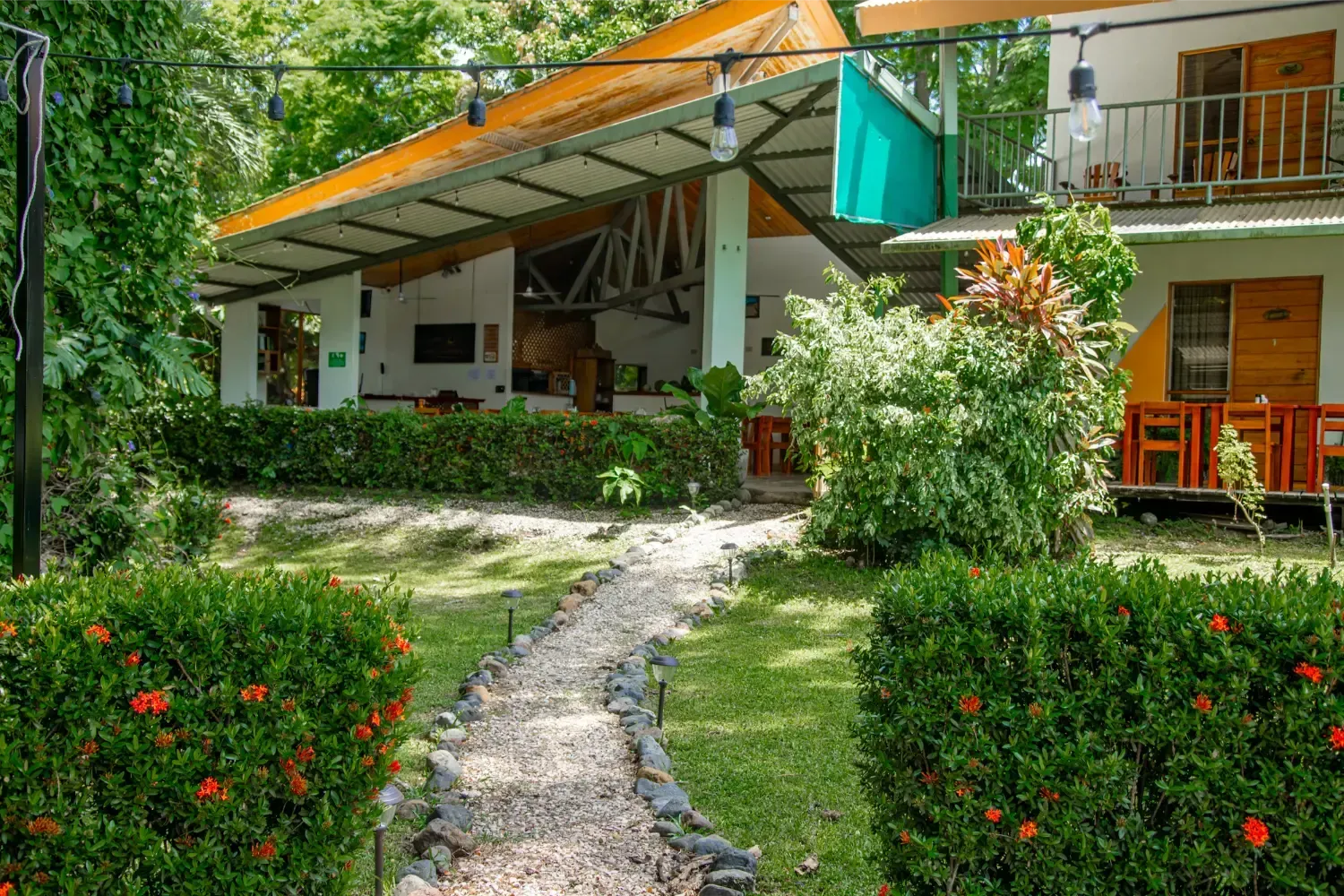 Stone path leads to a tropical lodge with a white facade, orange roof, and lush greenery.
