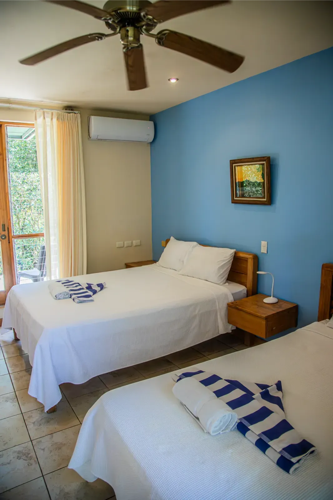 Bedroom with two beds, blue wall, open door to balcony. White bedding, blue and white striped towels.