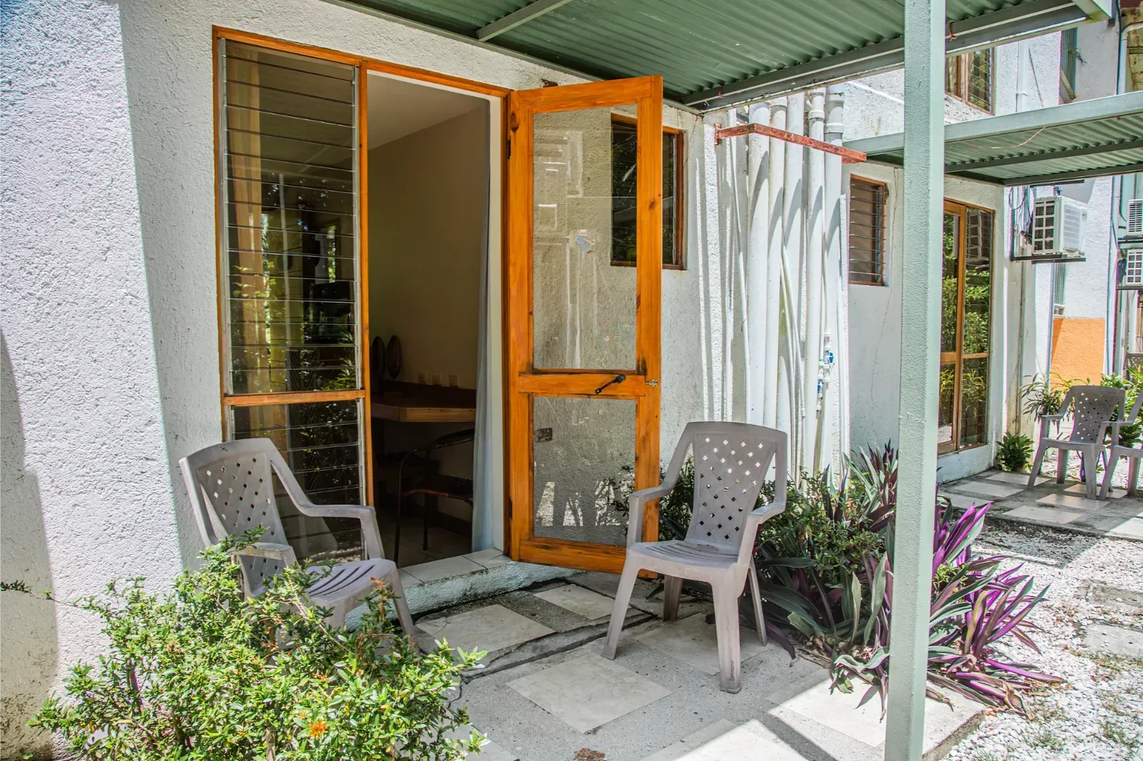 Exterior of a small white building with open wooden door, two chairs, and plants on patio.