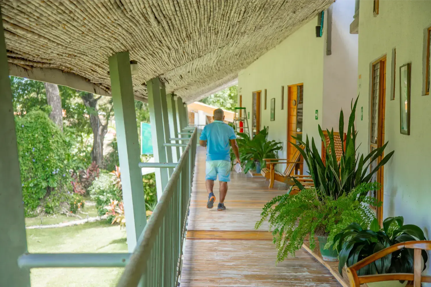 Man walking on a wooden balcony with plants and thatched roof; green landscape in the background.
