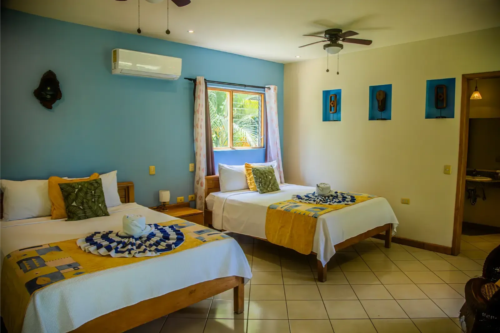 Bedroom with two beds, blue and yellow accents, window, air conditioner, artwork, and open door.