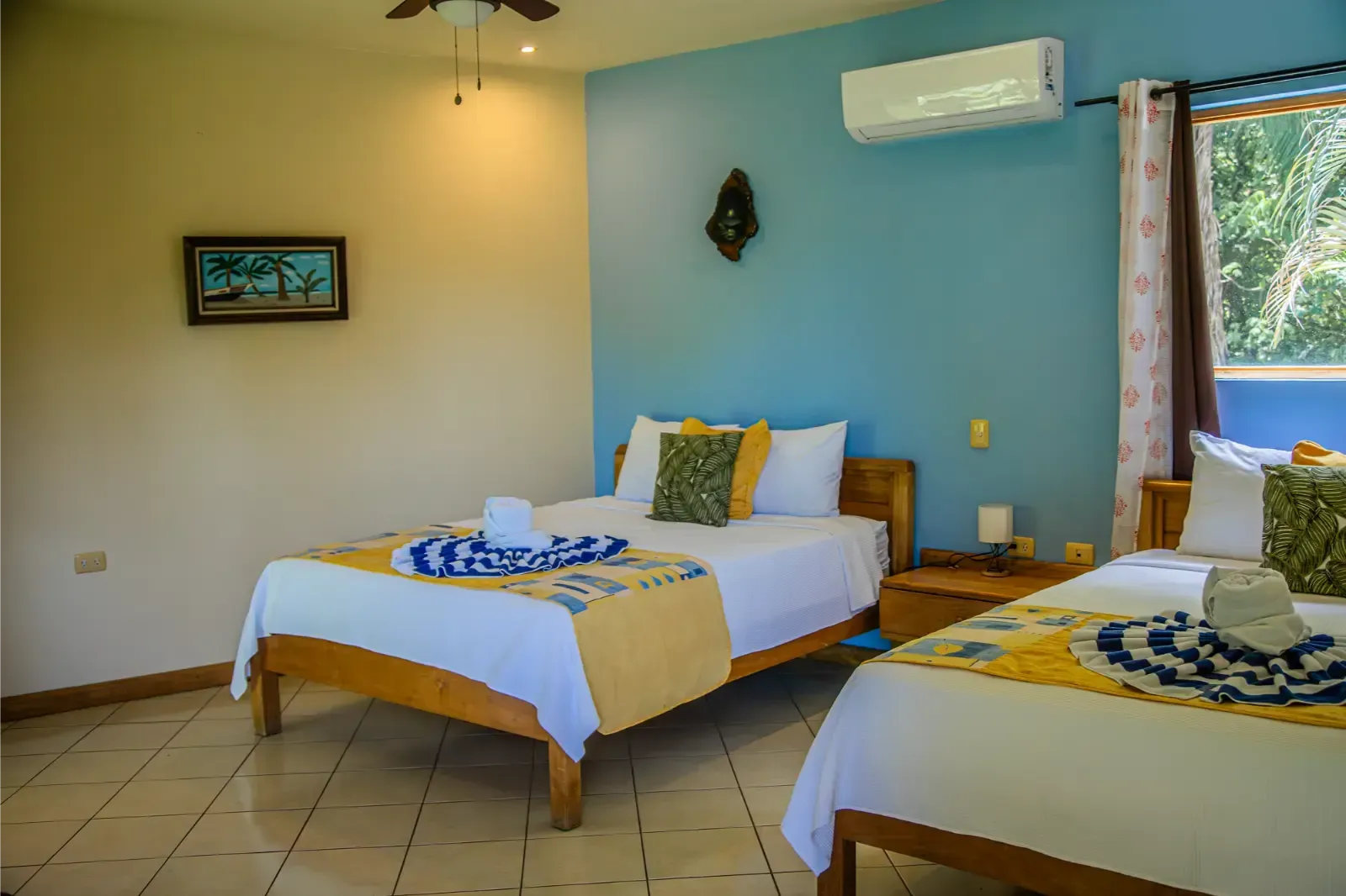 Two beds in a room with blue and yellow accents, air conditioning, and a window with greenery.