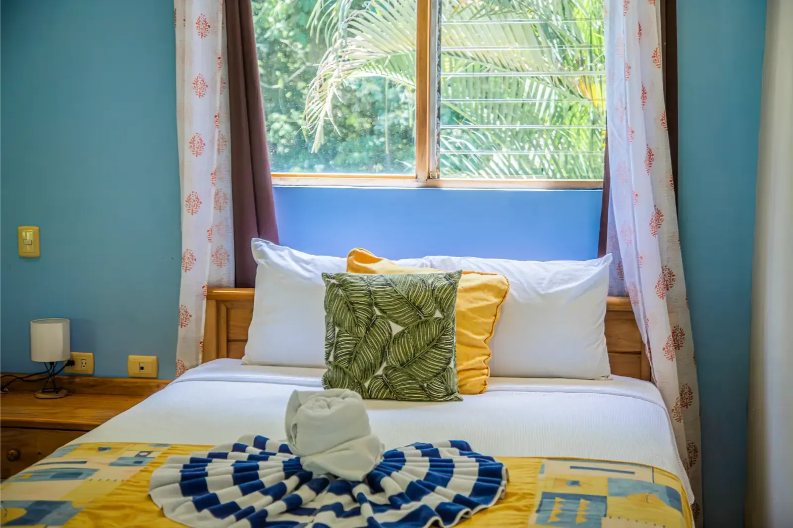 Bedroom with a bed, blue wall, window with greenery, hat, yellow blanket, and pillows.