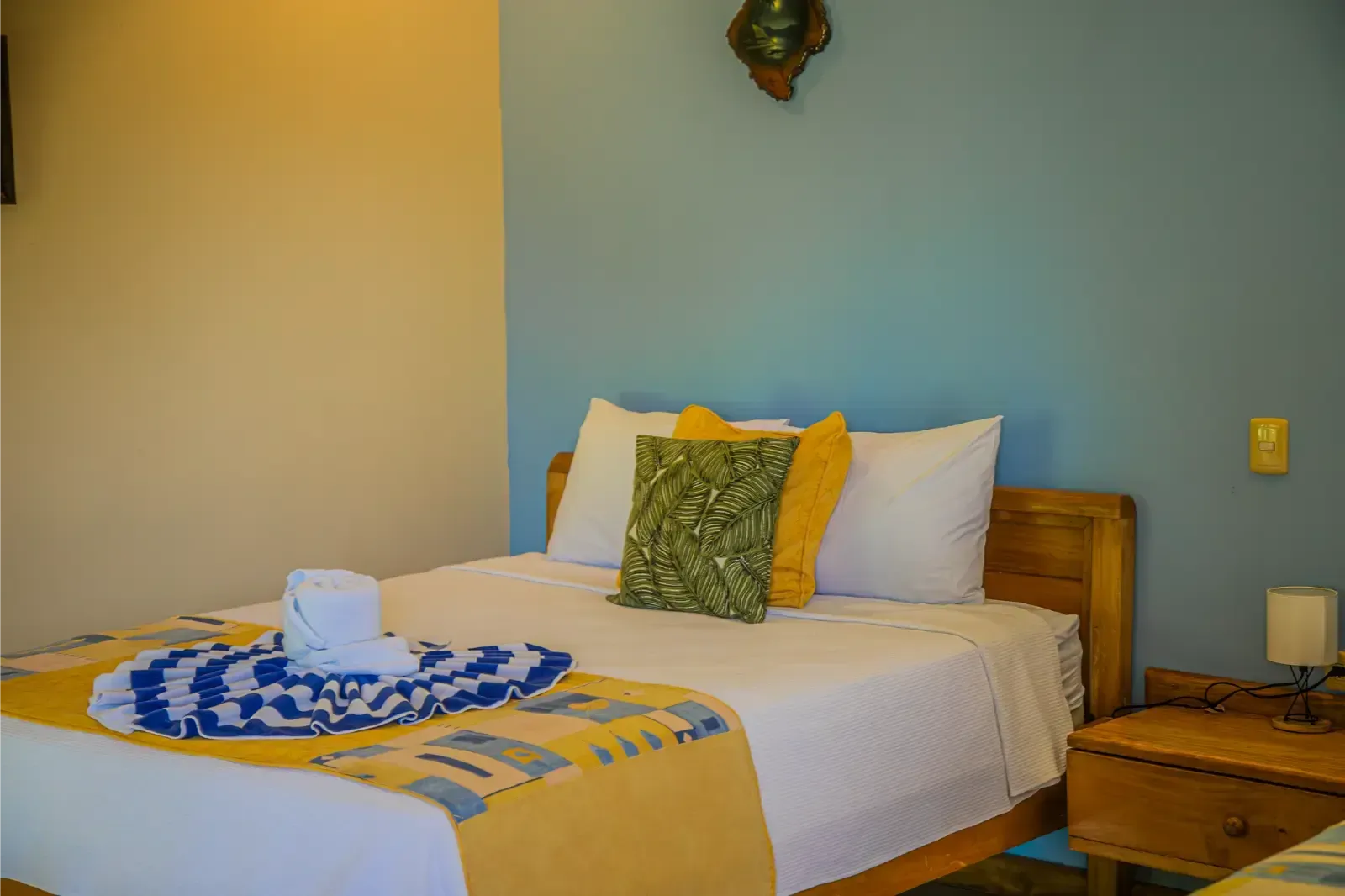 Bedroom with double bed, yellow and blue accents, wooden furniture, blue wall, light and dark yellow throw, towels.