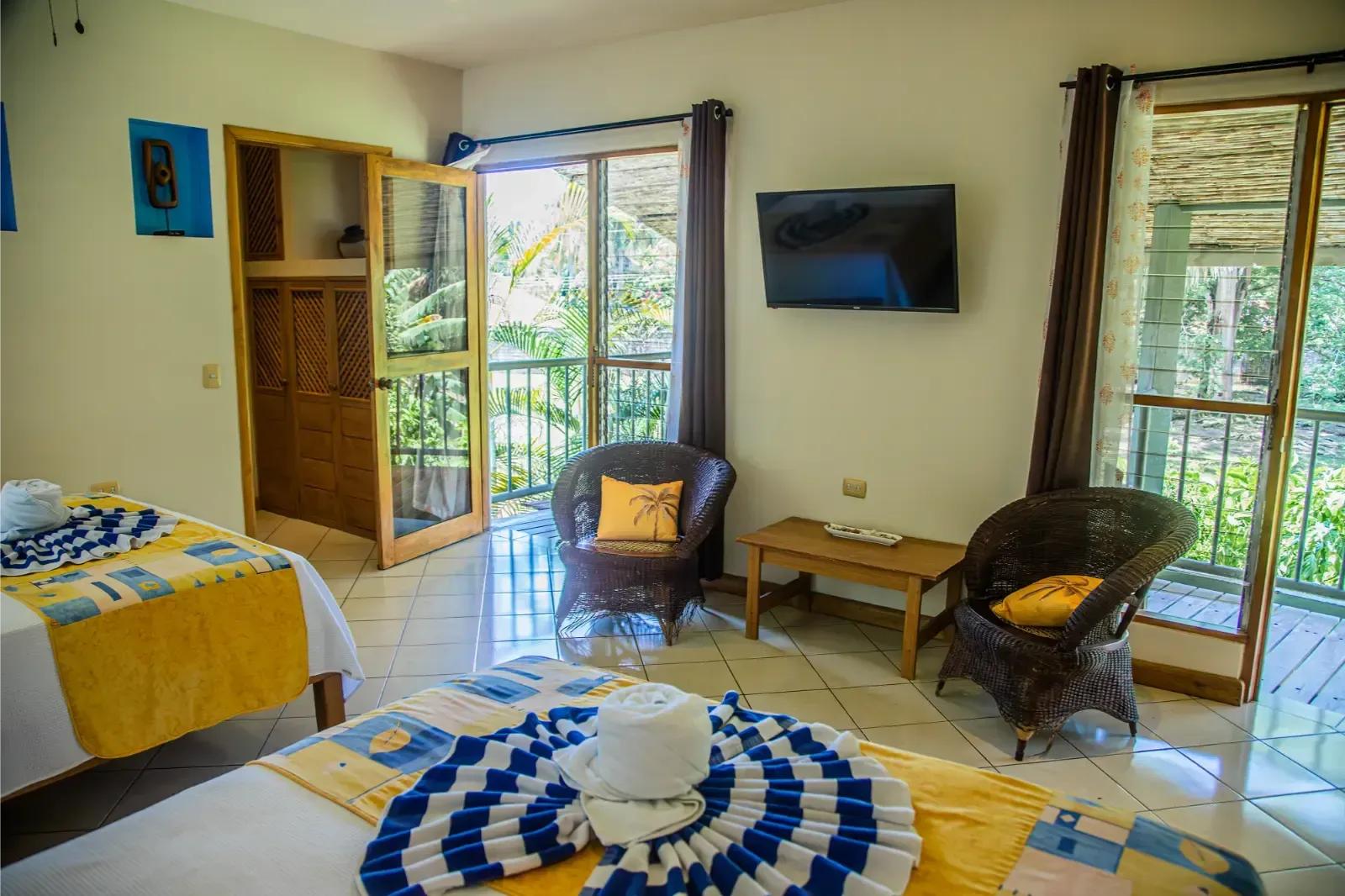Hotel room with two beds, balcony access, two wicker chairs, and a wall-mounted TV.