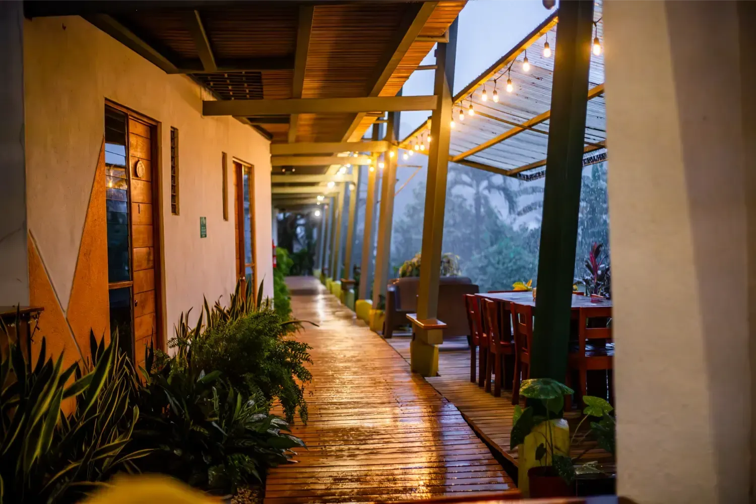Covered walkway with string lights, rain-slicked wood, tables, and plants; evening setting.