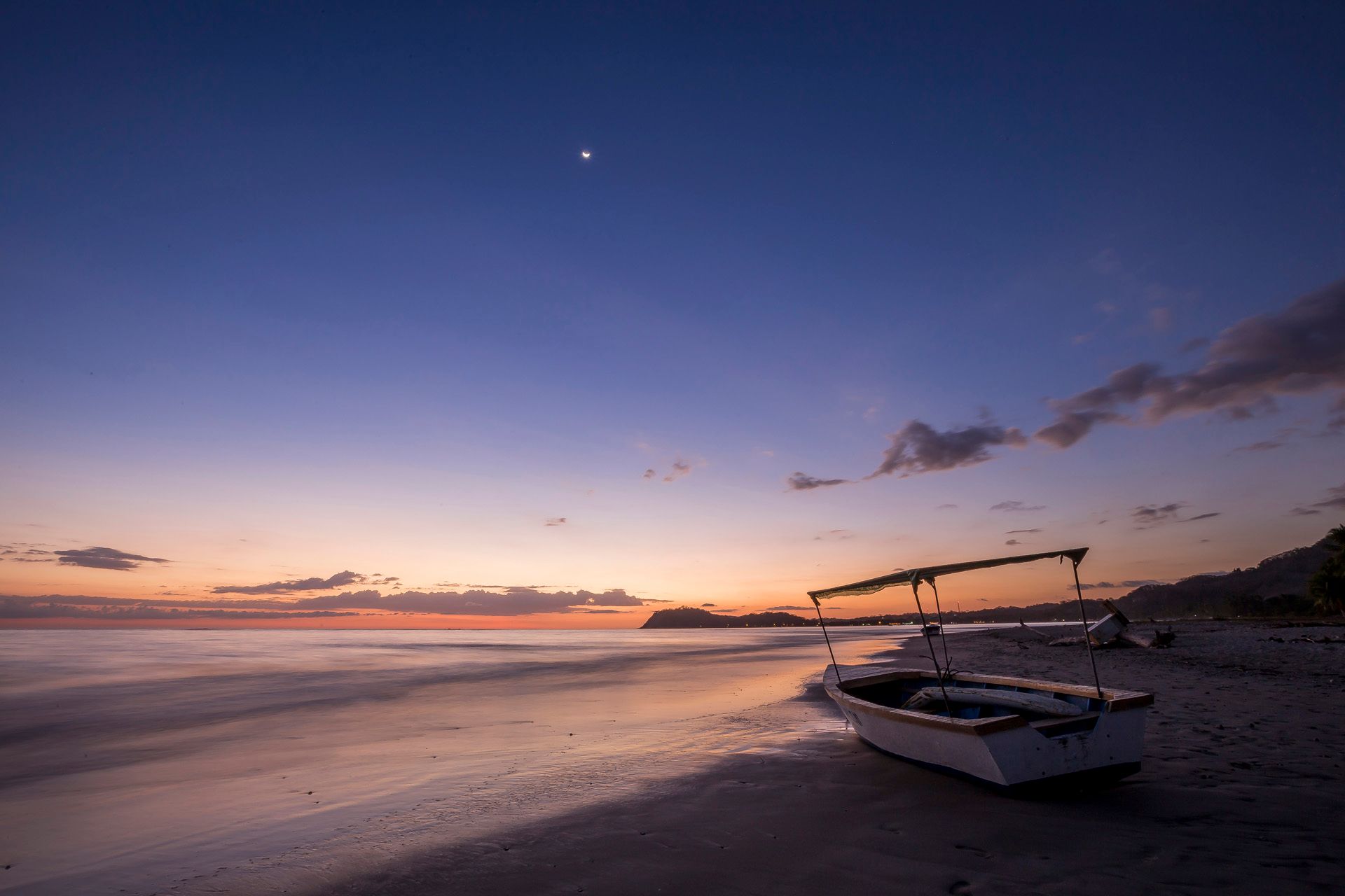 A boat rests on a beach at dawn, with a crescent moon in a blue and orange sky.