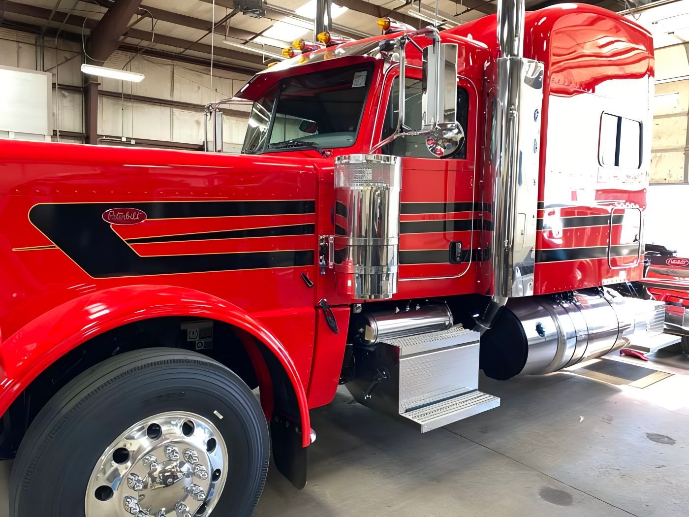 Red semi-truck with chrome accents, parked inside a brightly lit garage. Black stripes adorn the cab.