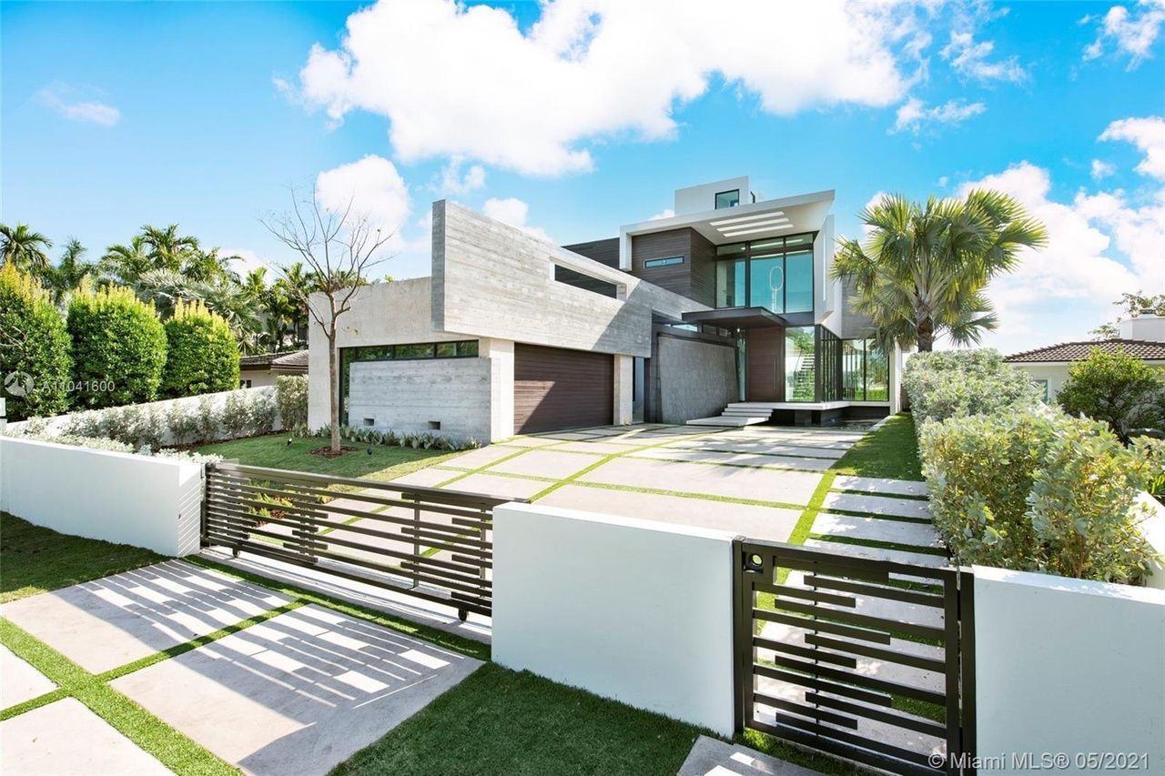 Modern two-story house with stone facade, glass entrance, driveway, and gated lawn on a sunny day.