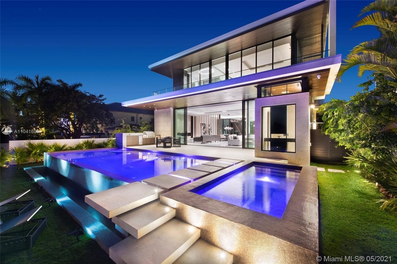 Modern luxury home with illuminated pools, outdoor seating, and lush landscaping at dusk.