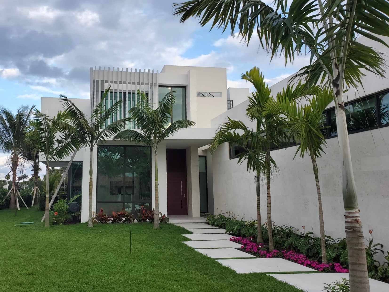 Modern white house with a walkway, palm trees, and green lawn under a cloudy sky.