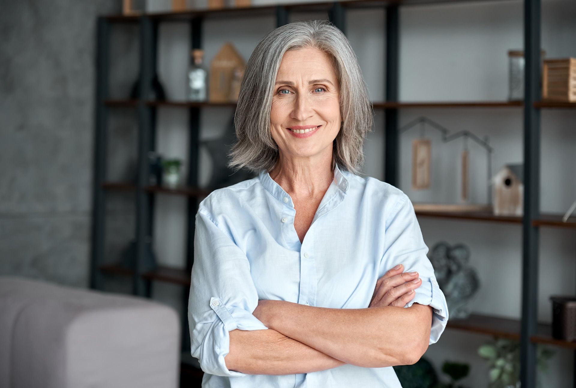 A woman with gray hair is standing in a living room with her arms crossed and smiling.