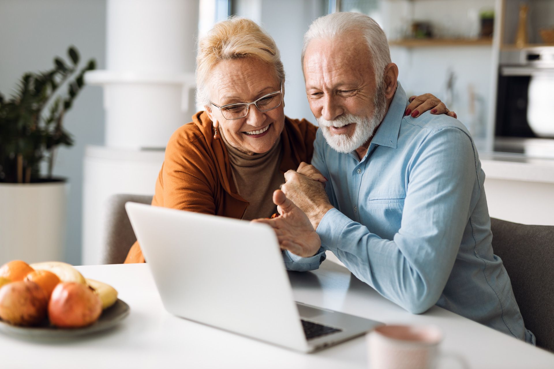 An elderly couple is sitting at a table looking at a laptop computer.