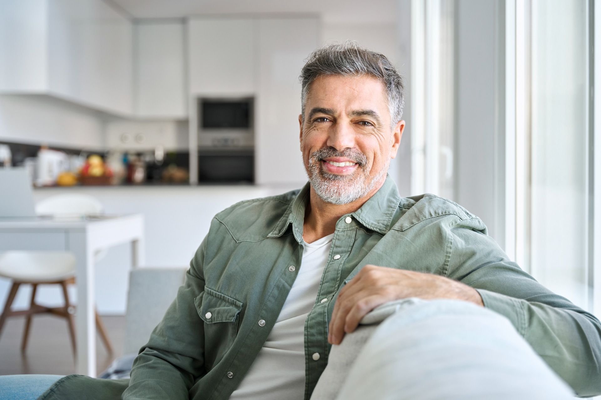 A man is sitting on a couch in front of a window and smiling.