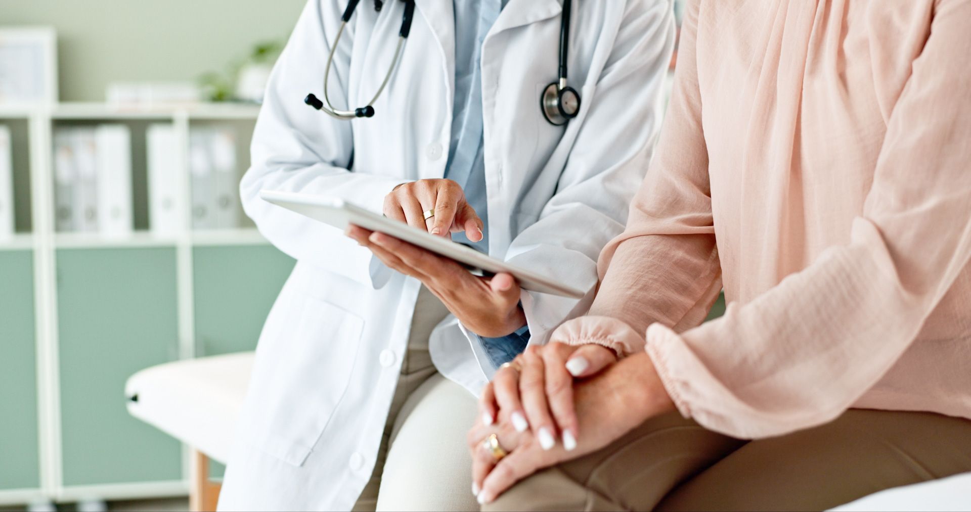 A doctor is holding the hand of a patient while looking at a tablet.