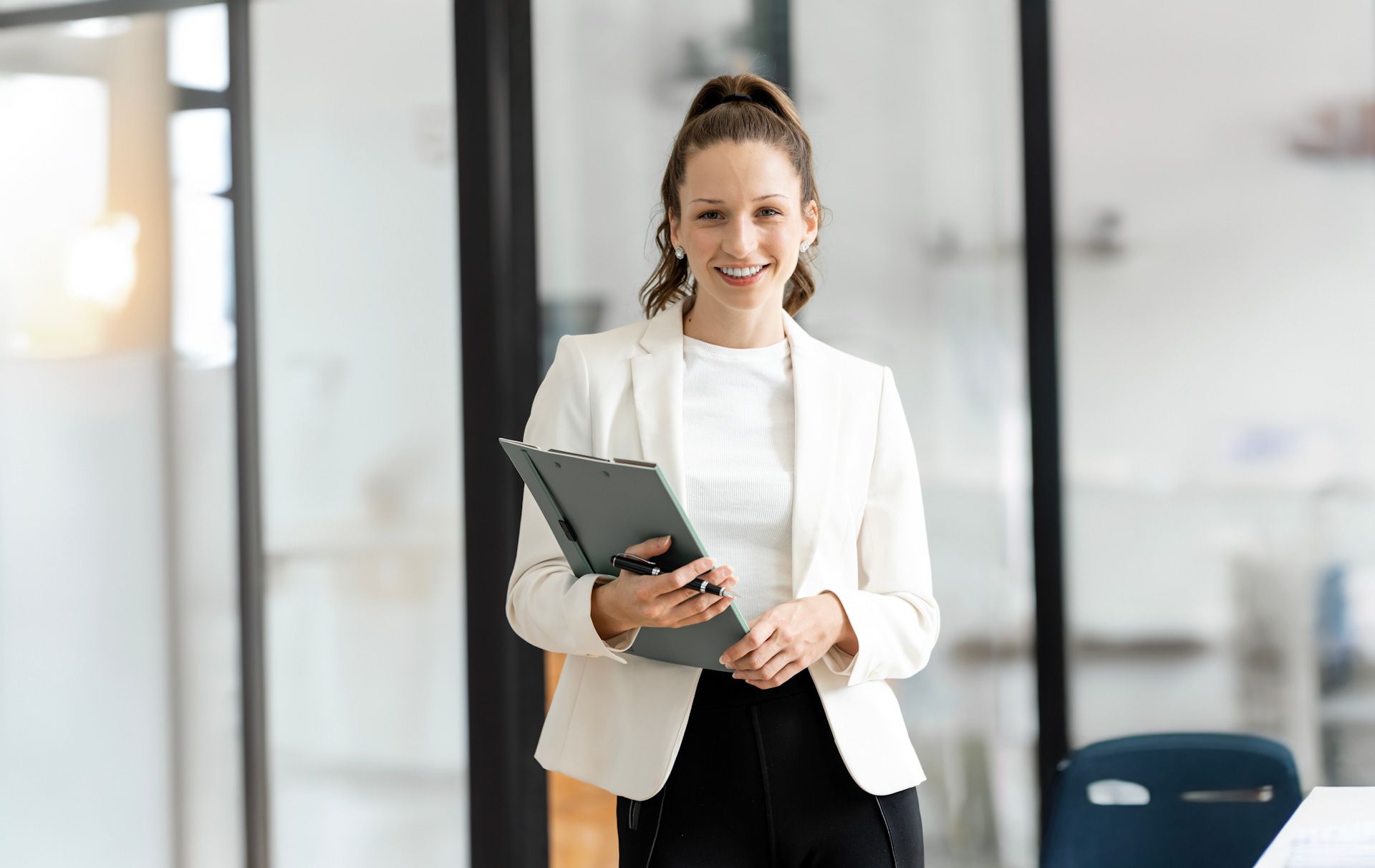 A woman in a white jacket is holding a clipboard and smiling.