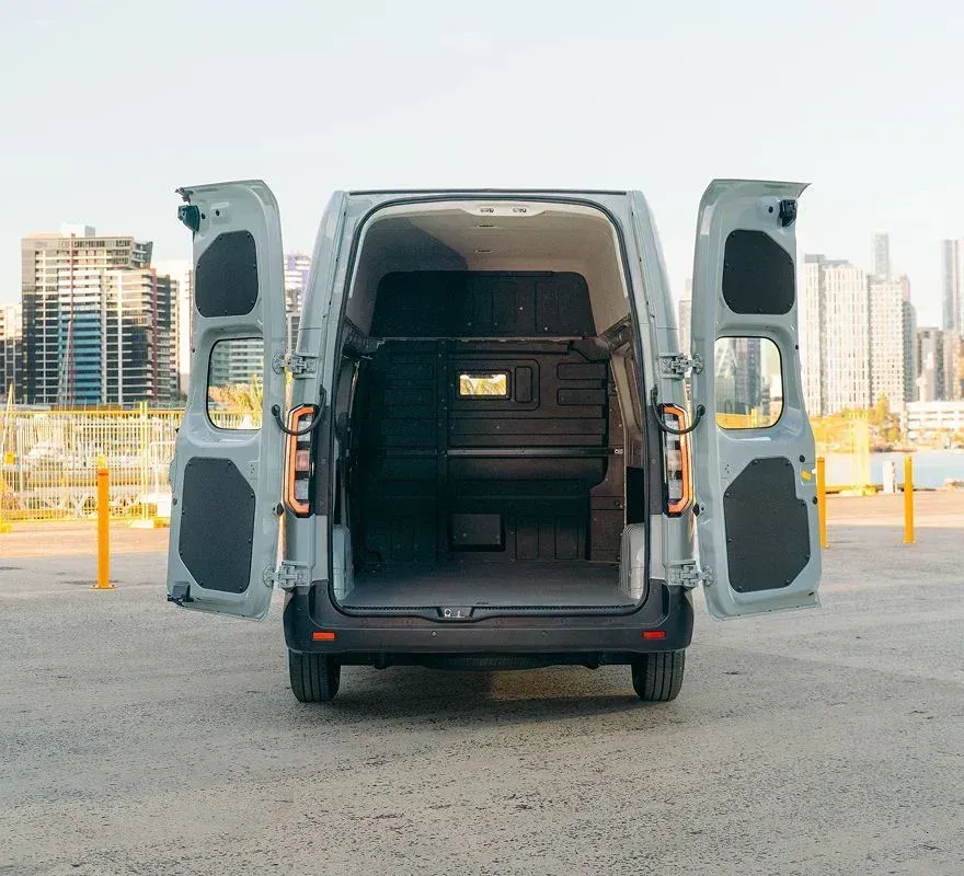 Rear view of a light gray van with open back doors, parked on an urban lot with city buildings in the background.