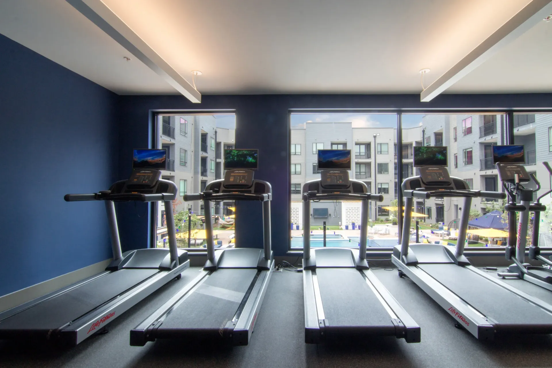 Gym interior with four treadmills lined up by large windows overlooking the outdoor pool.