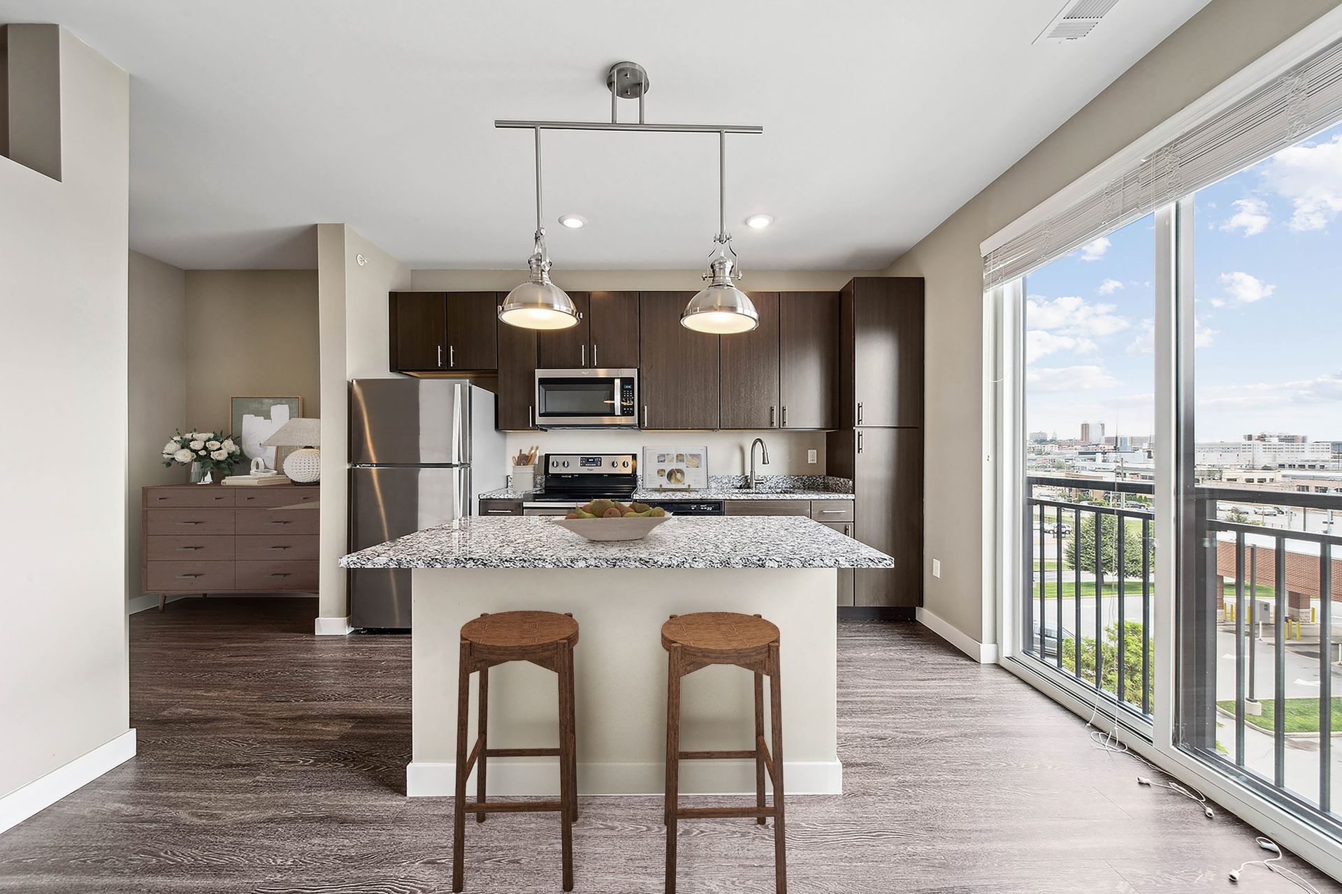 Modern apartment kitchen with granite island, stainless appliances, dark cabinets, and large balcony windows.