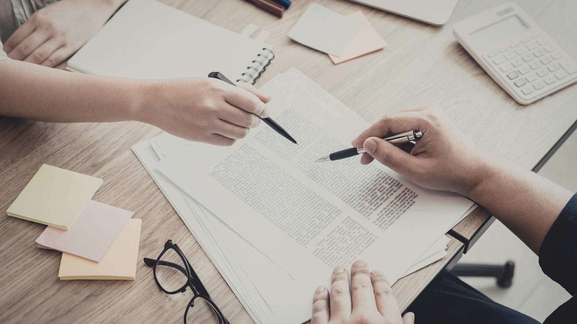 Hands reviewing a document at a desk with pens, notebooks, and glasses.