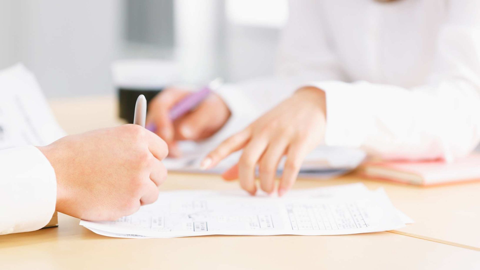 Hands writing on a document at a bright desk with a pen and papers