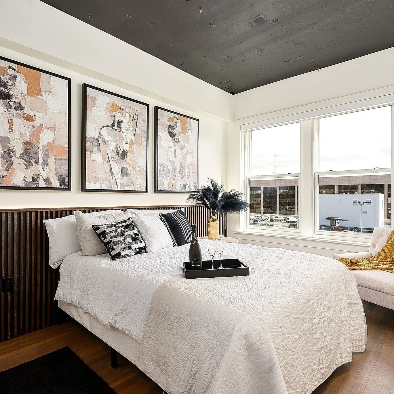 Bedroom with bed, artwork, and window. Cream bedding, black accents, gold blanket, and dark wood headboard.