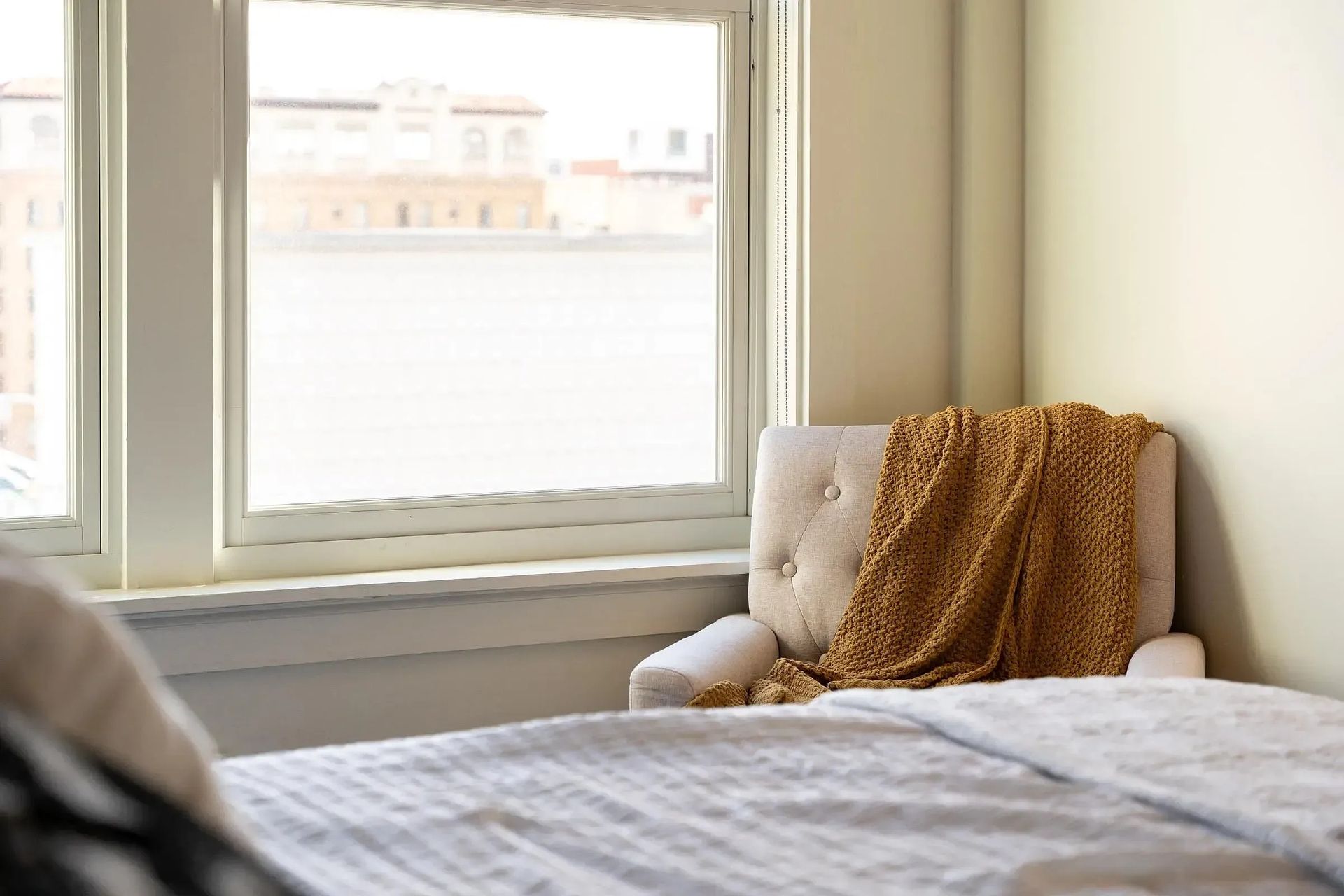 Cozy bedroom corner with a chair, blanket, and window overlooking a cityscape.