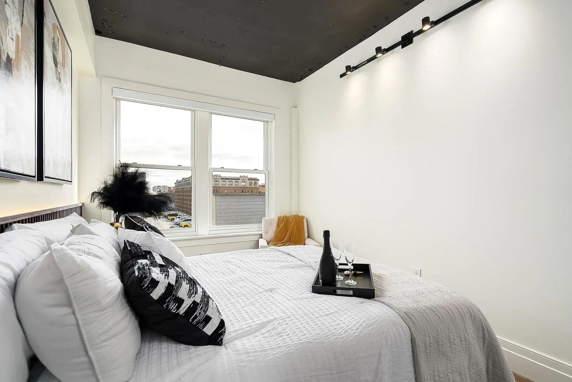 Bedroom with white bedding, black accent pillows, window seat, cityscape view, and black ceiling.