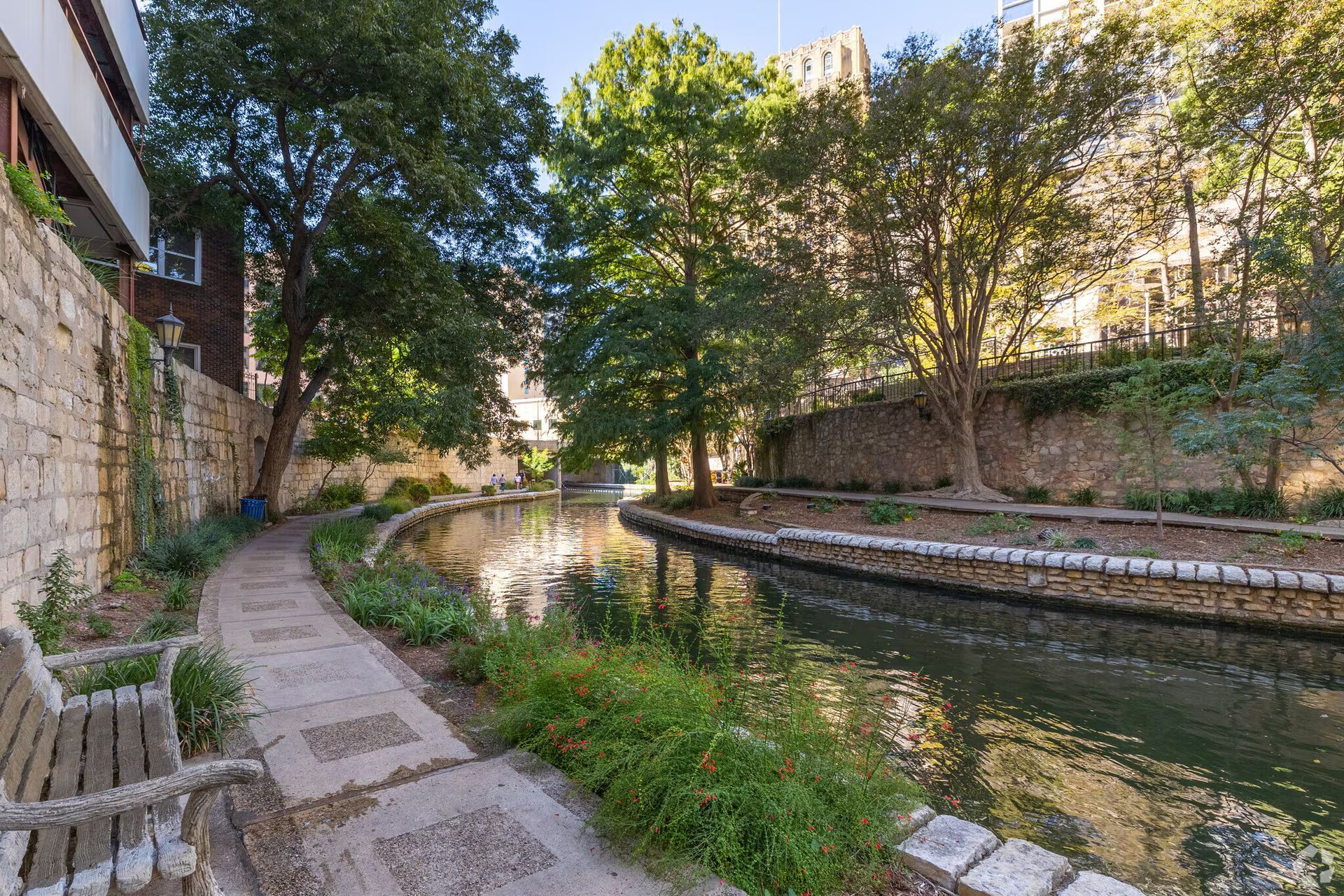 Riverwalk in San Antonio, TX, with stone walkways, lush greenery, and buildings lining the water.