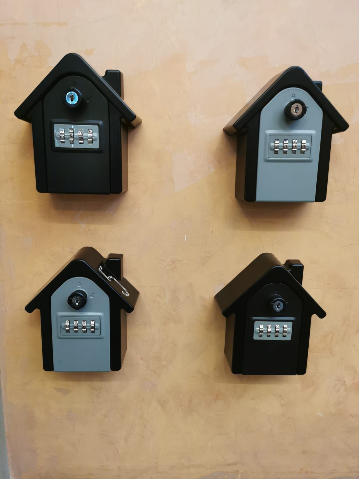 Four small wall-mounted house-shaped lock boxes in black, gray, and beige on a tan wall.