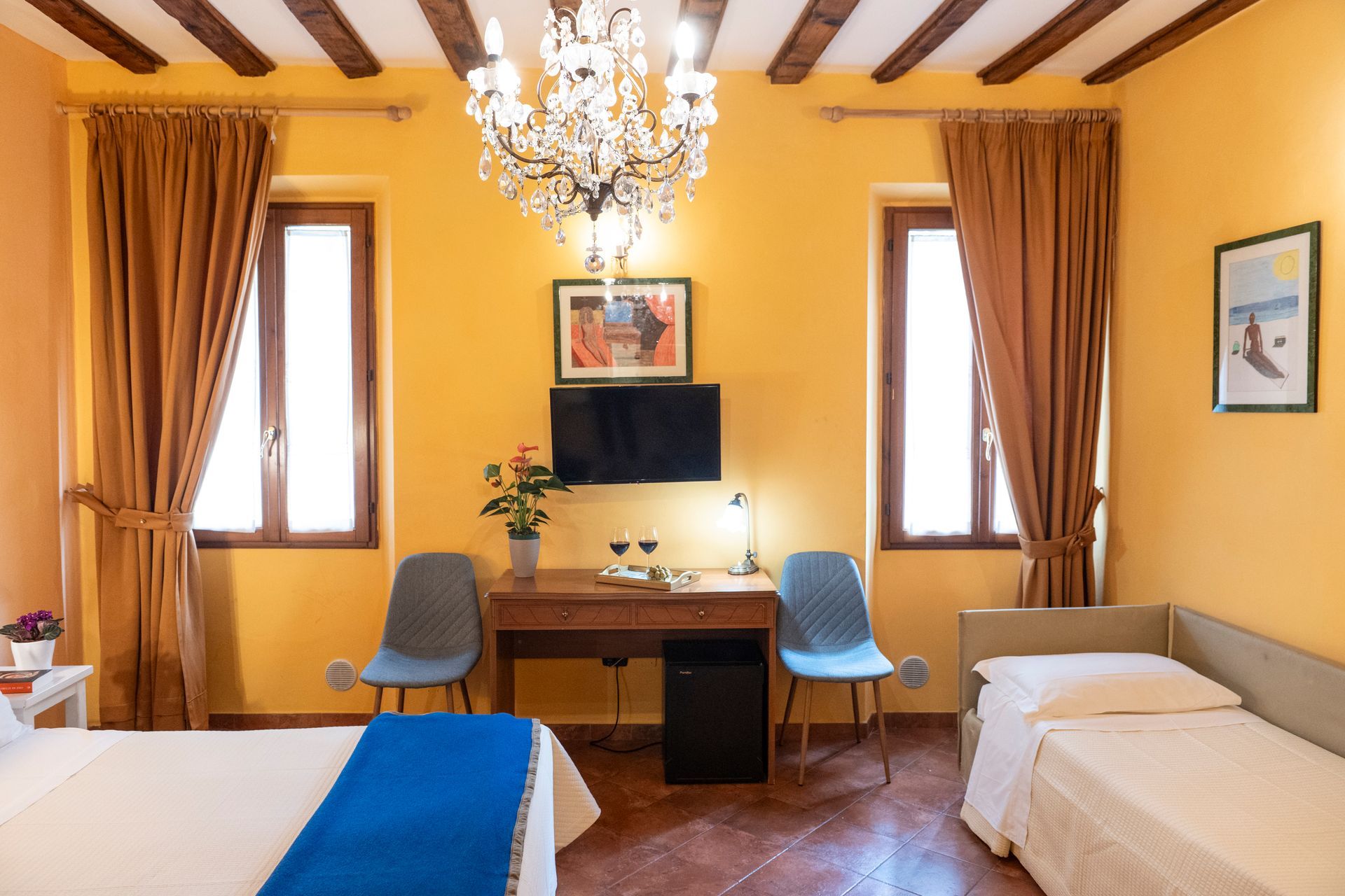 Bright yellow hotel room with twin beds, desk, chairs, TV, chandelier, and tall windows with curtains