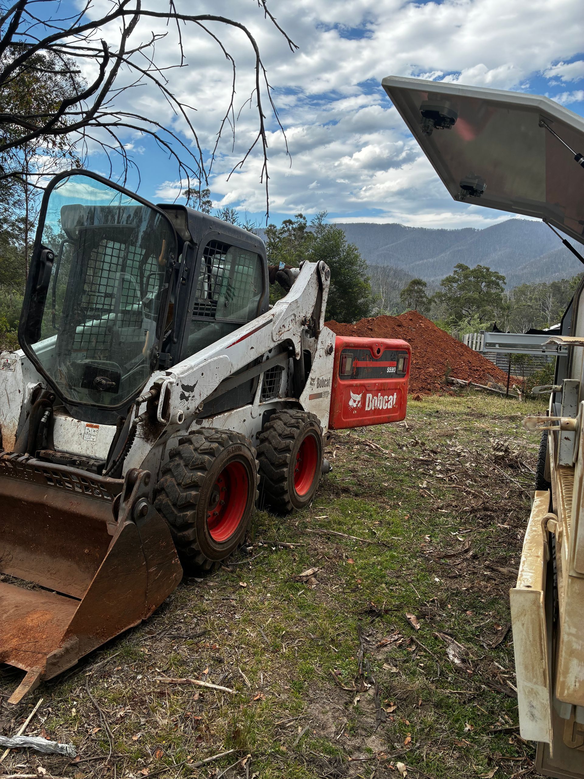 A white Bobcat skid-steer loader parked on grass, with an open bucket. Red wheels, open door, and a pile of red bricks in the background. — Bodes All Mechanical In Merimbula, NSW