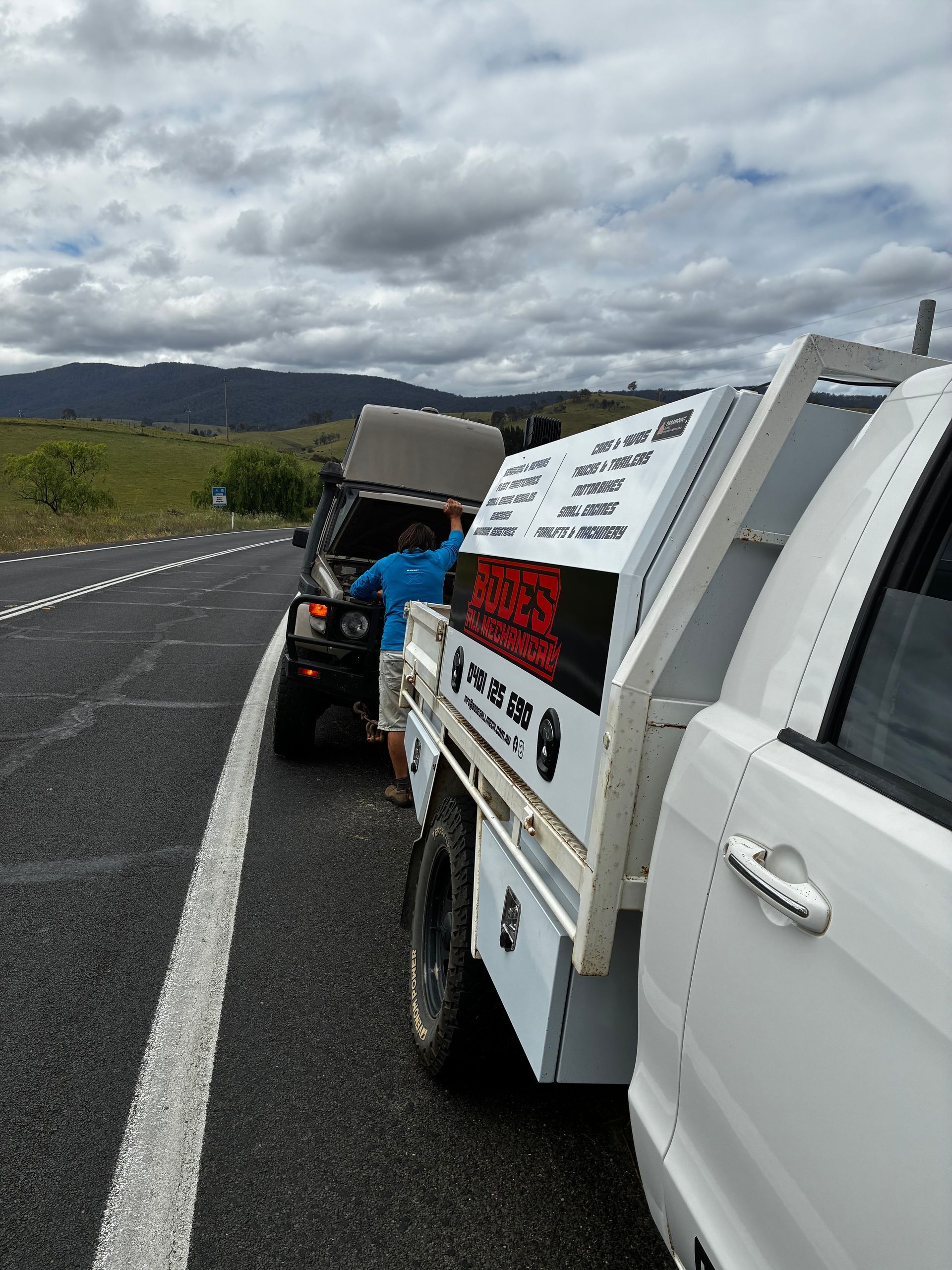 A white truck with a service body is parked on the side of a road, next to a black SUV with its hood up; a person is working. — Bodes All Mechanical In Tantawangalo, NSW