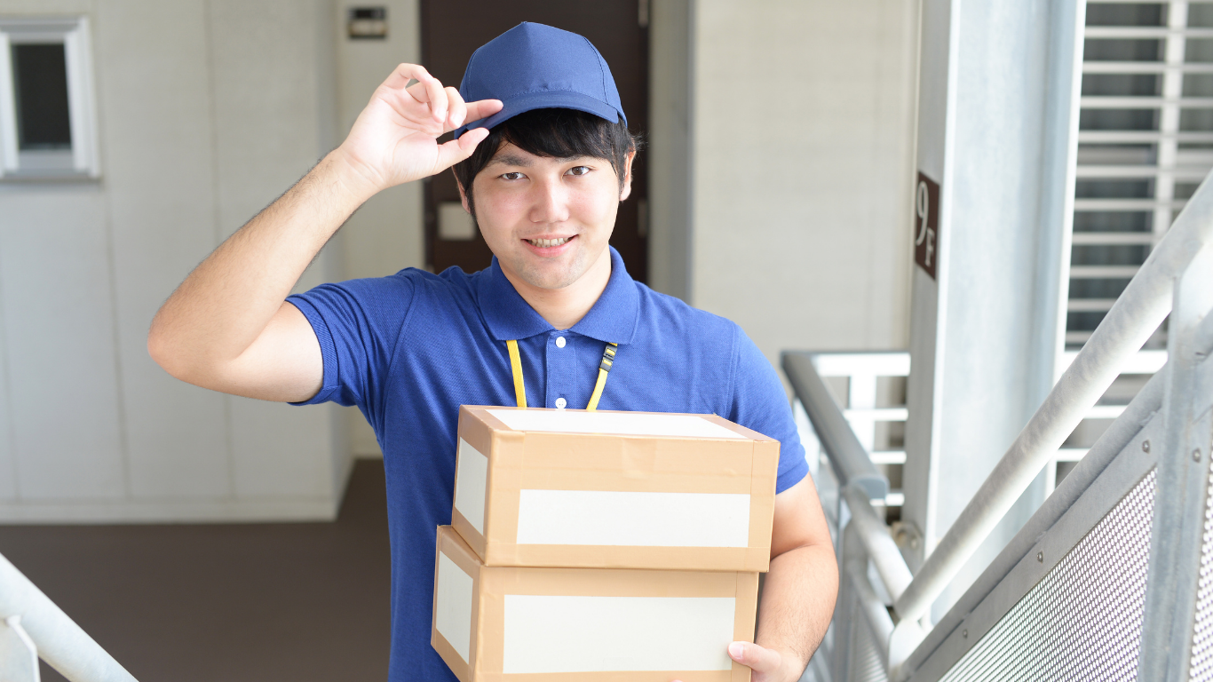 Delivery person in blue uniform holding boxes, tipping cap, smiling outdoors.