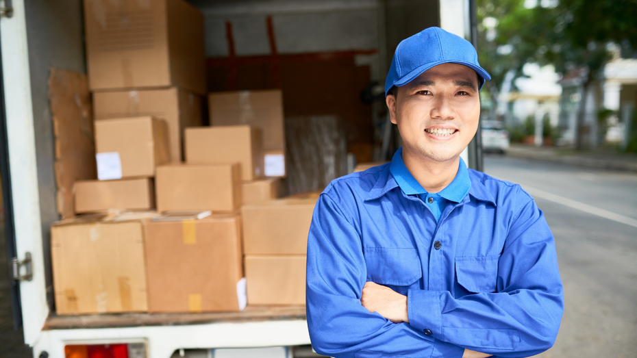 Delivery worker in blue uniform smiles, arms crossed, in front of a truck loaded with cardboard boxes.