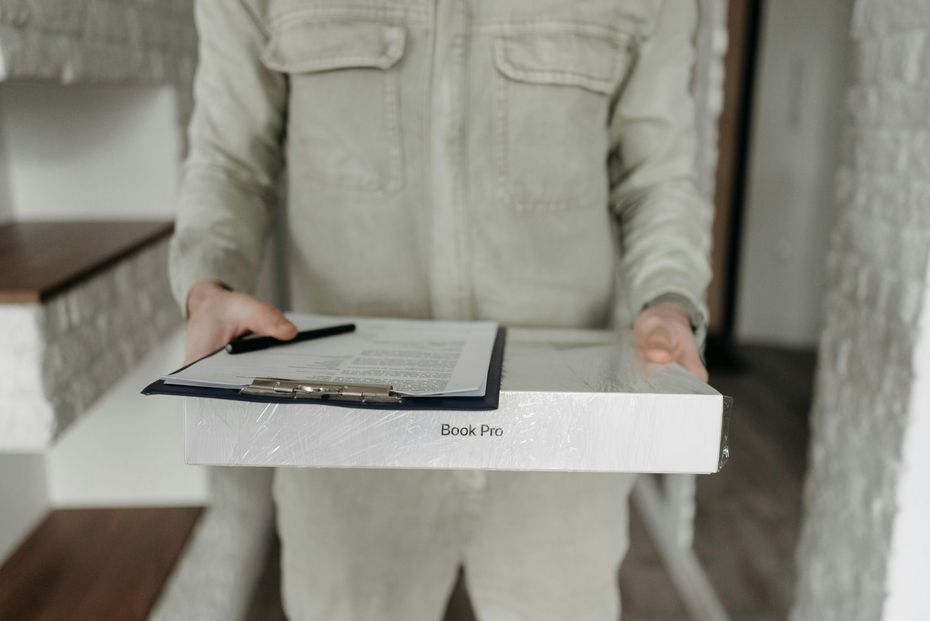 Person in beige jumpsuit holding a clipboard and a delivery box, standing indoors.