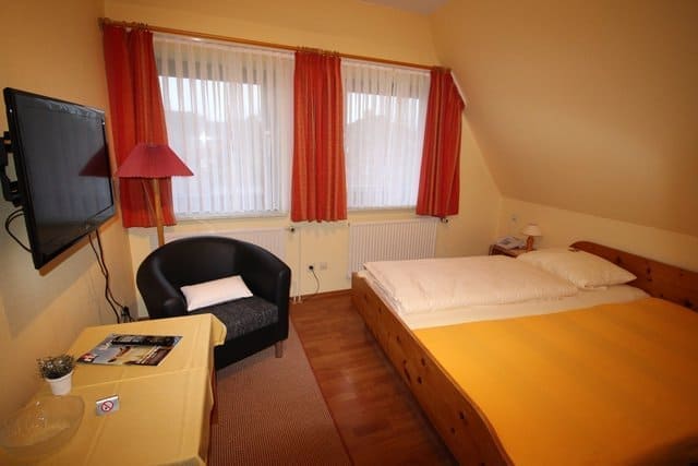 Small bedroom with a double bed, armchair, TV, and a window with red curtains. The room is lit with warm colors.