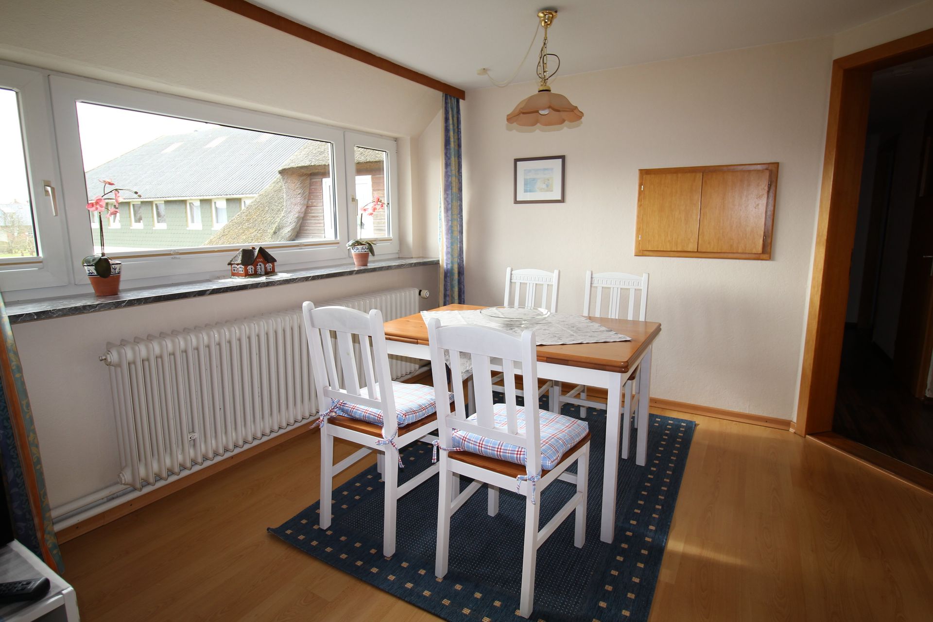 A dining room with a wooden table, four white chairs, a blue rug, and a window. Sunlight streams in.