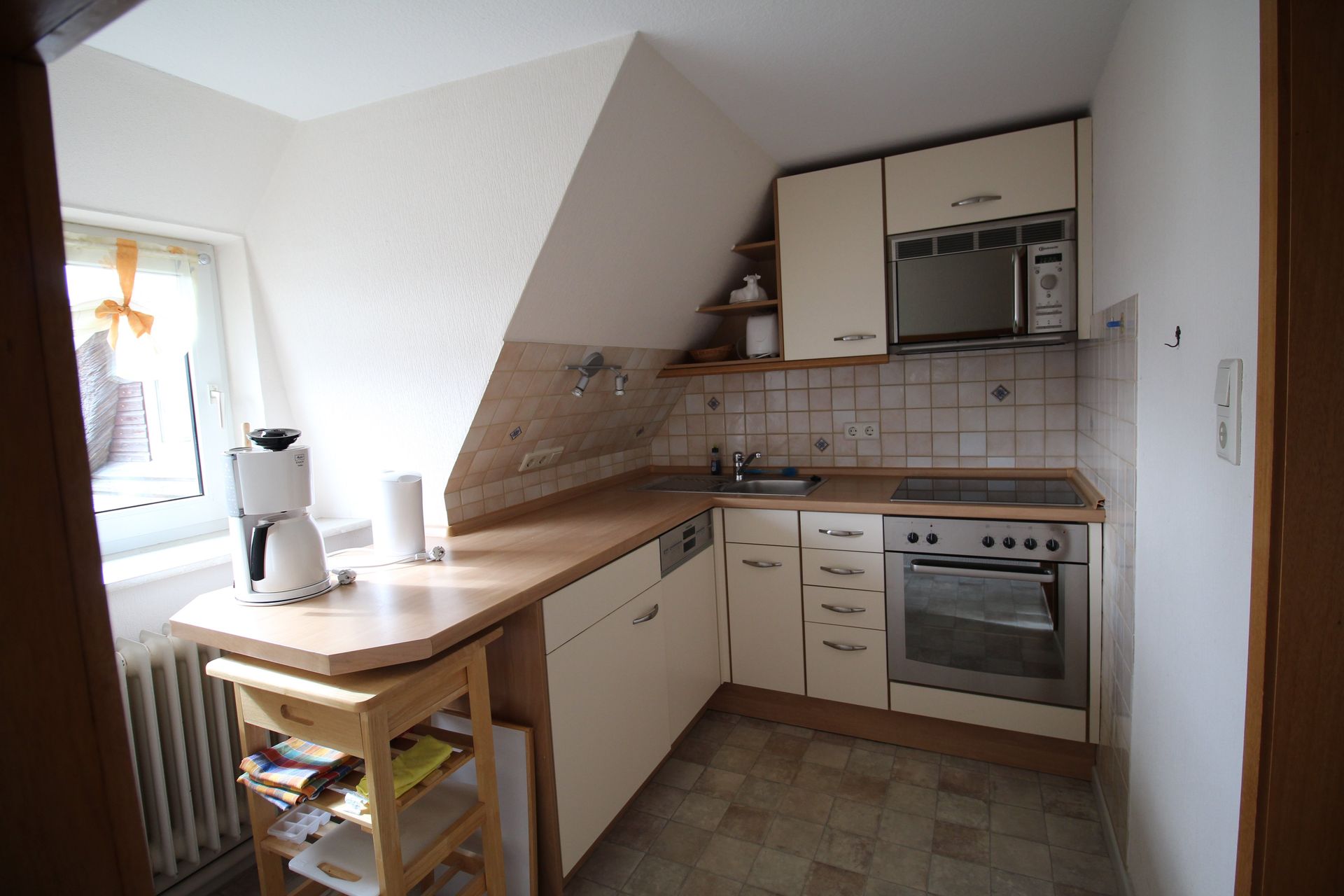 Small kitchen with angled ceiling, featuring a stove, microwave, cabinets, and small table near a window.