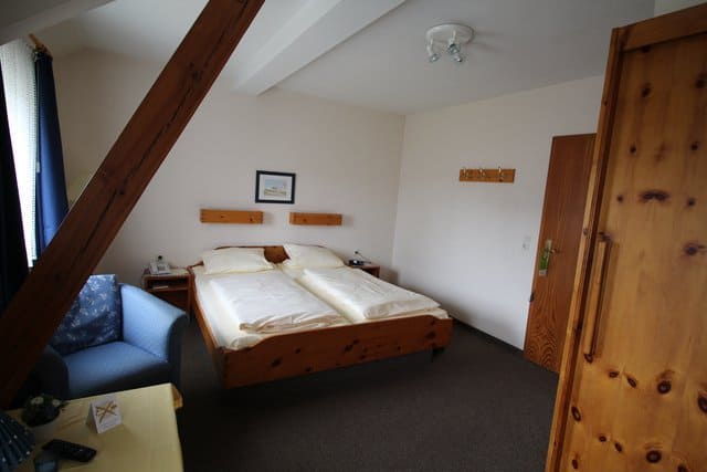 Bedroom with a bed, blue armchair, wooden beams, and a wooden door. Neutral colors with white walls and a dark carpet.