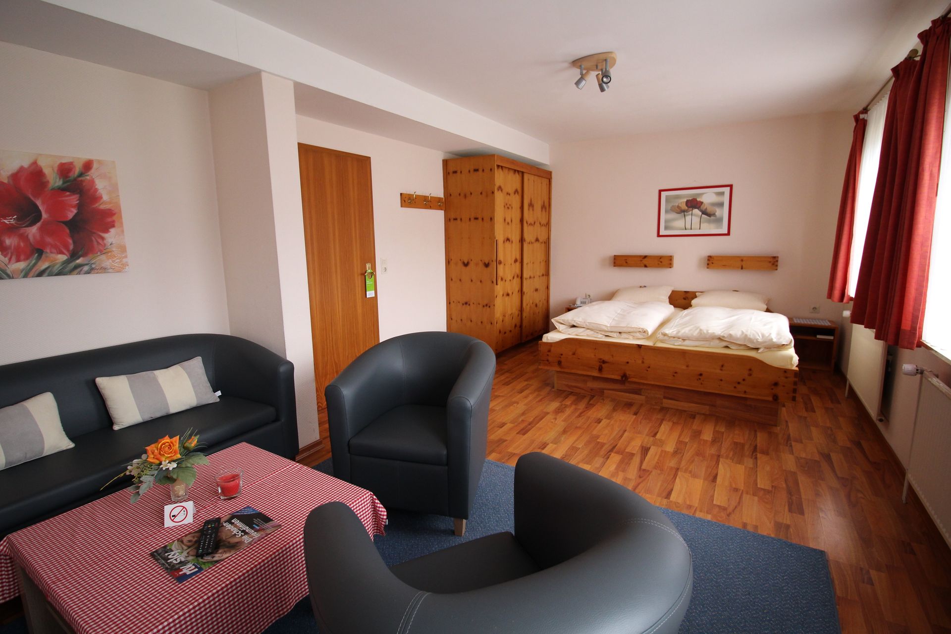 A hotel room with a couch, chairs, bed, wooden furniture, red curtains, and artwork.