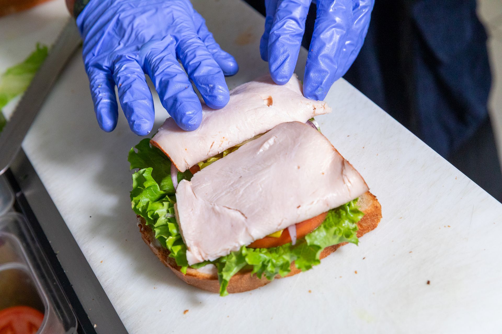 Hands in blue gloves assembling a sandwich with turkey, lettuce, and tomato on a white cutting board.