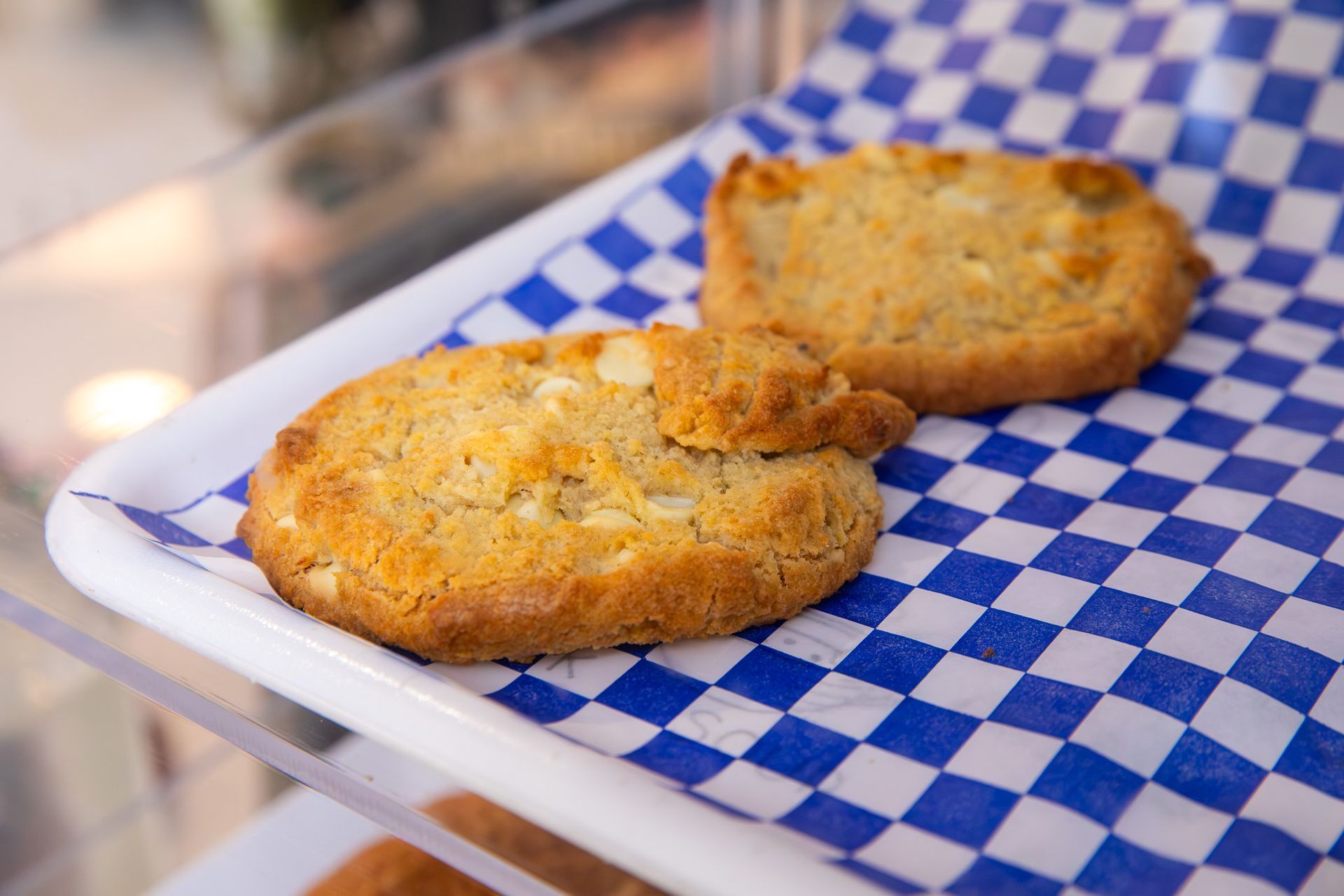 Two golden-brown cookies with white chocolate chips on blue and white checkered paper.