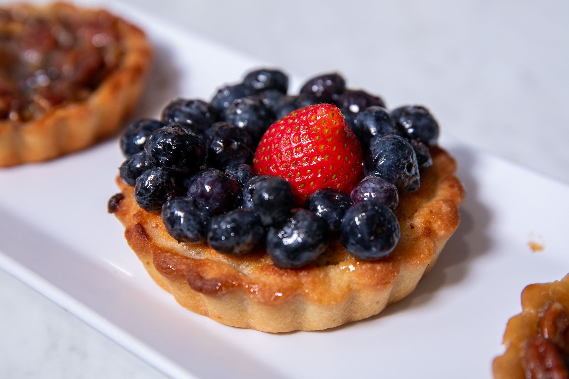 Blueberry tart with a strawberry on a white rectangular plate.