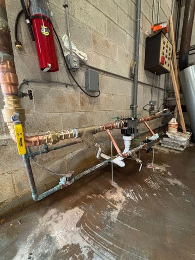 Basement utility area with exposed pipes, valves, and a red fire extinguisher on a concrete wall.