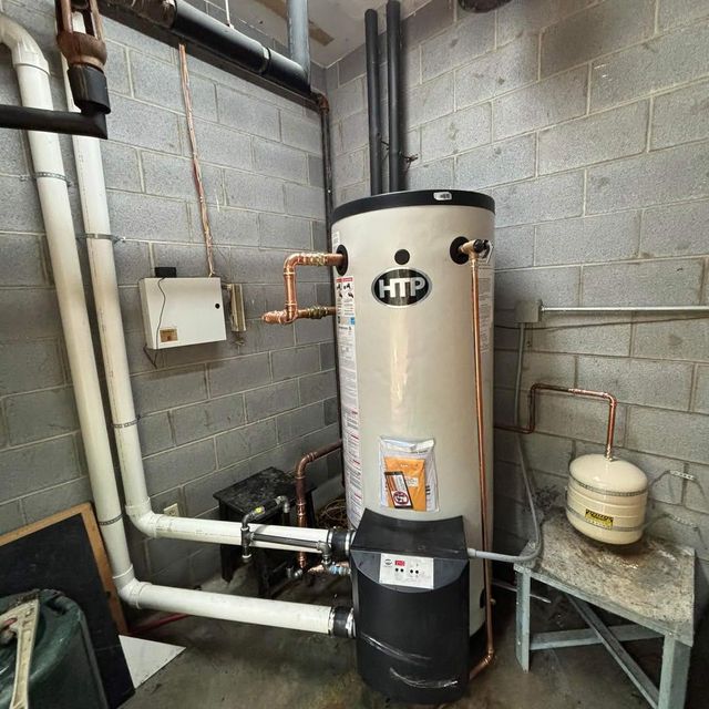 Water heater in a basement utility room beside pipes, cinderblock walls, and a small tank on a table