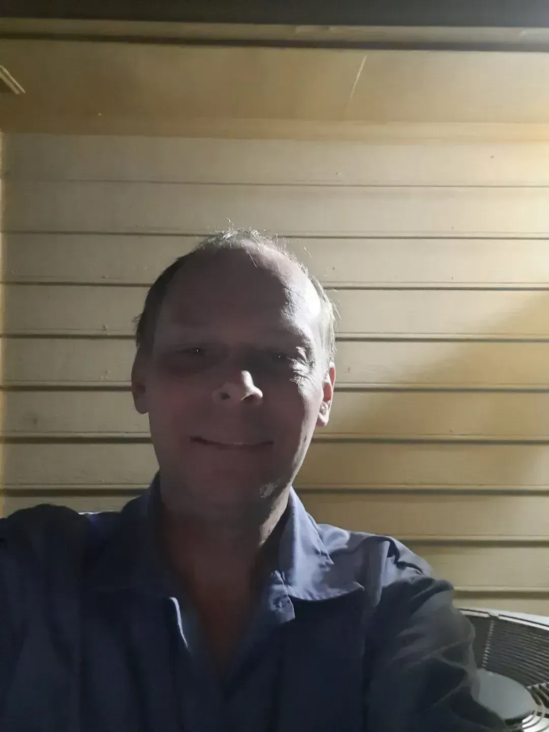 Man smiling, wearing a blue shirt, in front of a tan, horizontally-lined wall.