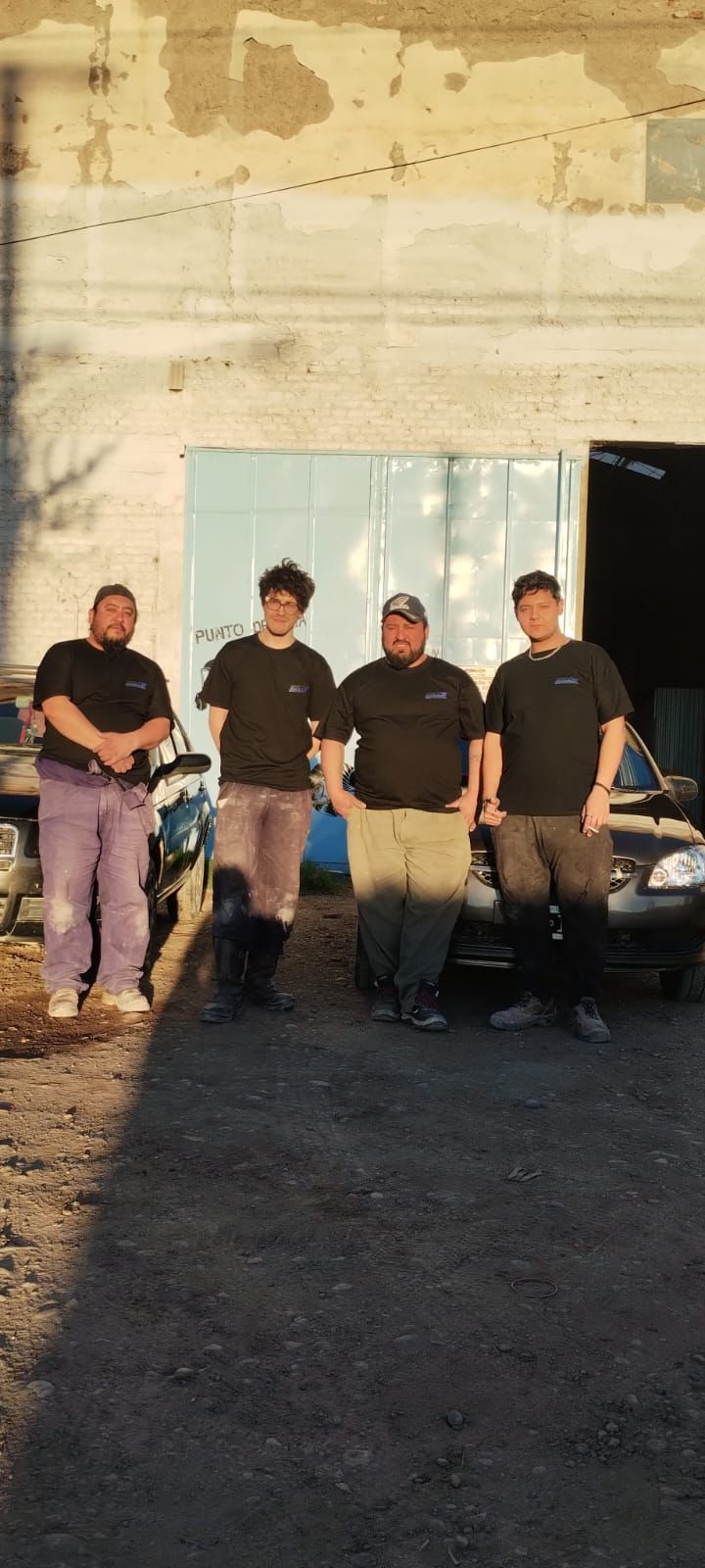 Cuatro personas, todas con camisa negra, frente a un garaje. Dos coches aparcados cerca.