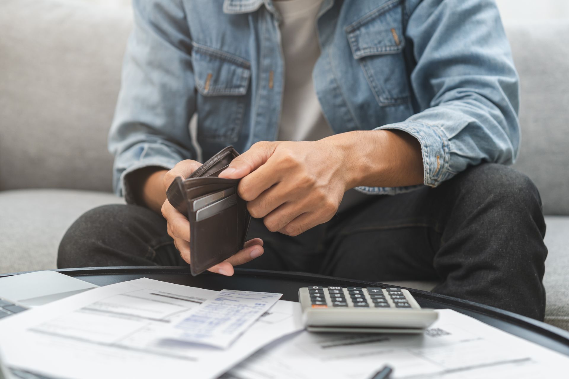 Man sitting on a couch, holding an empty wallet, looking at bills and a calculator.