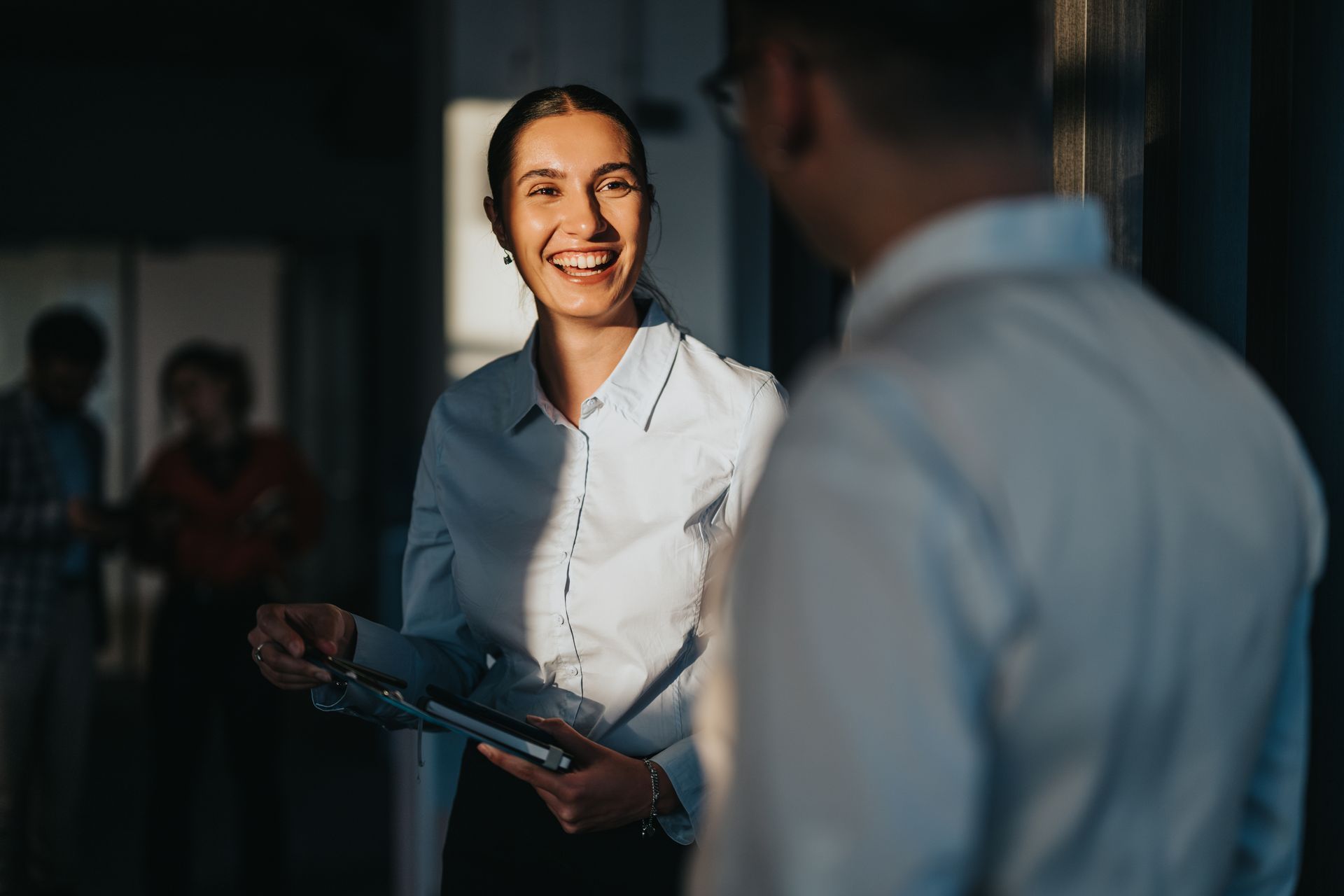 Woman in white shirt smiles at a man, holding a clipboard, in an office setting.